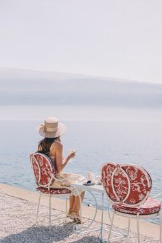 a woman sitting at a table with two red chairs next to the ocean and drinking tea