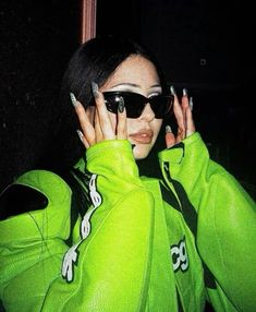 a woman with black nails and green hoodie holding her hands to her face while wearing sunglasses