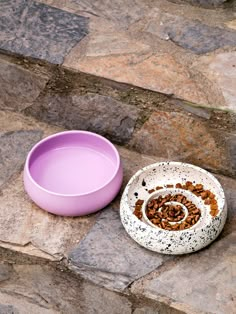 a dog bowl with food in it sitting next to a pink bowl on the ground