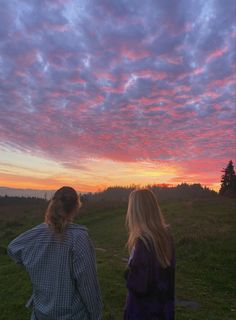 two women are sitting on the grass watching the sun go down with pink clouds in the sky