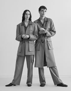 two people standing next to each other in front of a white wall wearing coats and slacks