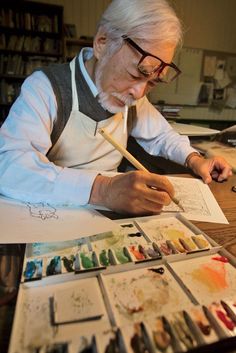 an older man sitting at a table working on some art work with watercolors