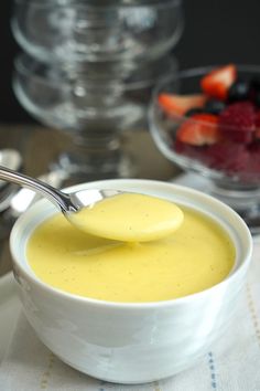 a spoon in a white bowl filled with yellow liquid and strawberries on the side