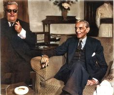 two men in suits are sitting on a couch and one is pointing at the camera