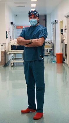 a man in scrubs and red shoes stands with his arms crossed wearing a surgical mask