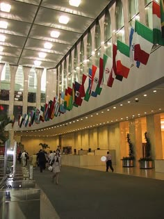 people are walking through an airport lobby with many flags hanging from the ceiling above them