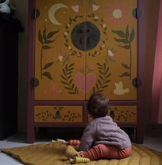 a small child sitting on the floor in front of a cabinet with flowers painted on it