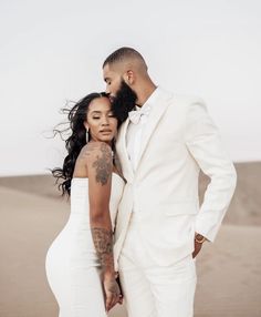 a man and woman standing next to each other in the desert with their arms around each other