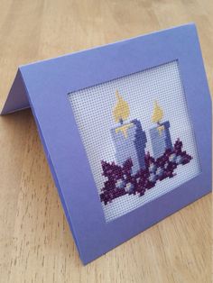 a cross - stitch picture frame with two candles in it on a wooden table next to a candle holder