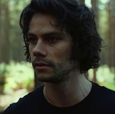 a man with long hair standing in the woods looking off into the distance while wearing a black t - shirt