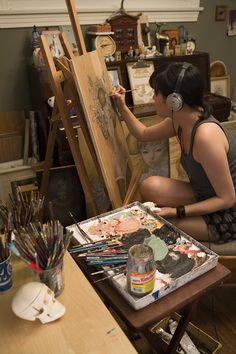 a woman sitting in front of an easel painting with headphones on her ears