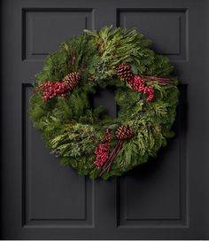 a green wreath with red berries and pine cones hanging on a black front door,