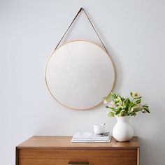 a white vase with flowers sitting on top of a wooden dresser next to a round mirror