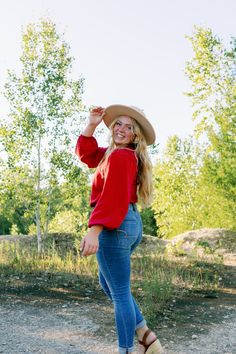 a woman in red shirt and cowboy hat standing on dirt road with trees behind her