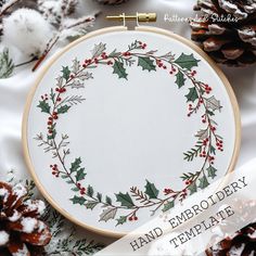 a hand embroidered christmas wreath with holly and pine cones on white fabric surrounded by snow