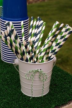 several striped paper straws in a bucket on top of green grass with blue and white cups behind them