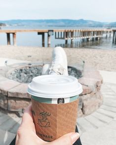 a person holding up a cup of coffee in front of the ocean and dock area