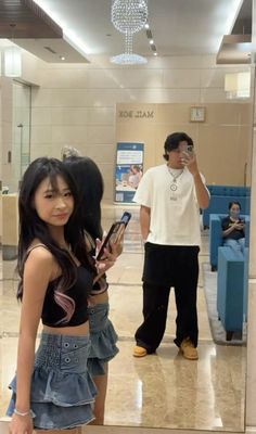 a girl standing in front of a mirror looking at her cell phone while another man looks on