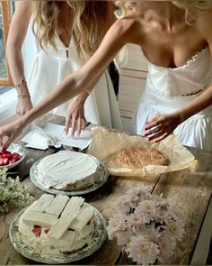 two women in white dresses standing over a table with cakes and flowers on it,