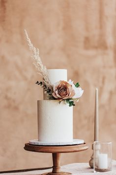 a white wedding cake with flowers and greenery sits on a wooden stand next to a candle