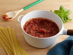 a white bowl filled with pasta and sauce next to some green leaves on a wooden table