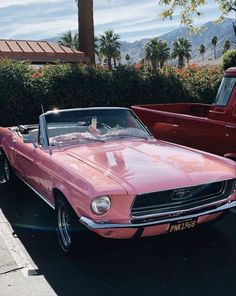 an old pink mustang convertible parked in a parking lot next to a red pickup truck