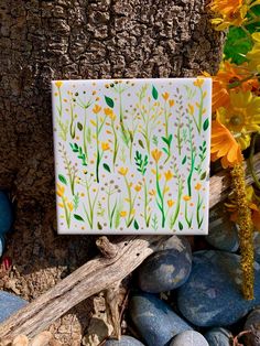 a white tile with yellow and green flowers sitting on rocks next to a tree trunk