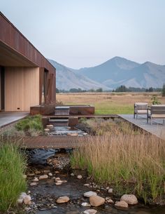 a wooden bench sitting next to a small stream in front of a building with mountains in the background