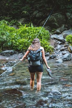 a woman walking across a river while holding onto a fishing rod and wearing a backpack