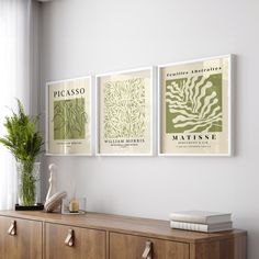 three framed posters on the wall above a dresser in a white room with a potted plant