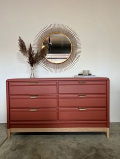 a red dresser sitting next to a round mirror on top of a wall with a plant in front of it