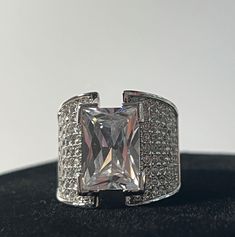 a white gold ring with a princess cut diamond surrounded by pave diamonds on a black surface