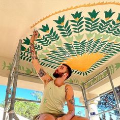 a man sitting on top of a stepladder painting a ceiling with green leaves