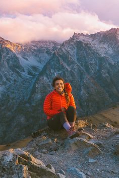 a woman in an orange jacket sitting on top of a rocky hill with mountains in the background