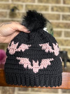 a hand holding a knitted batman hat with pink and black bats on the front