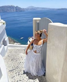 a woman in a white dress leaning against a wall with the ocean in the background