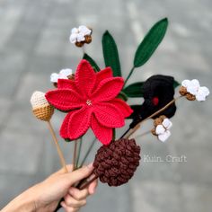 a hand holding a crocheted poinsettia with pine cones and flowers