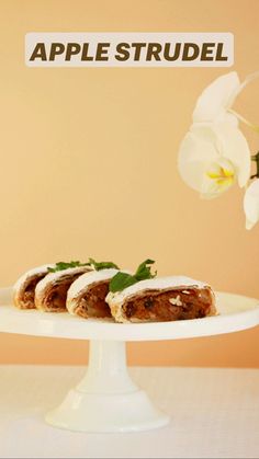 an apple strudel is sitting on a white cake plate with flowers in the background