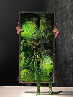 a person holding up a painting with green plants in the middle and two hands pointing at it