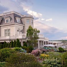 a large white house surrounded by lots of flowers and trees in front of it on a cloudy day