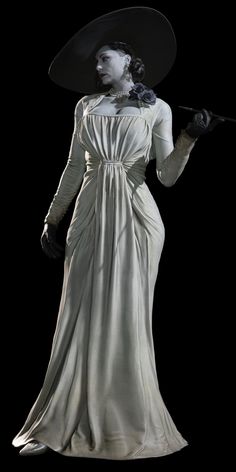 a woman in a long white dress and black hat holding a cane with both hands