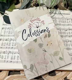 an open book sitting on top of sheet music next to some flowers and greenery