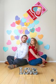 a man and woman sitting on the floor in front of a wall with paper hearts