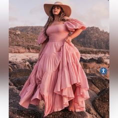 a woman in a pink dress and hat standing on rocks with her hands behind her back