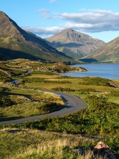 a winding road in the middle of a valley with mountains in the background and a body of water surrounded by grass