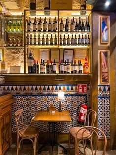 two wooden chairs sitting at a table in front of a wall with bottles on it