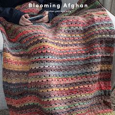 a woman sitting on a couch holding a cell phone in her hand while wearing a multicolored crocheted blanket