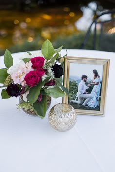 an arrangement of flowers and pictures on a white table cloth with a gold frame sitting next to it