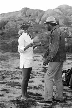 two men and a woman are standing in the desert talking to each other, with mountains in the background