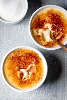 two white bowls filled with food sitting on top of a gray table next to spoons